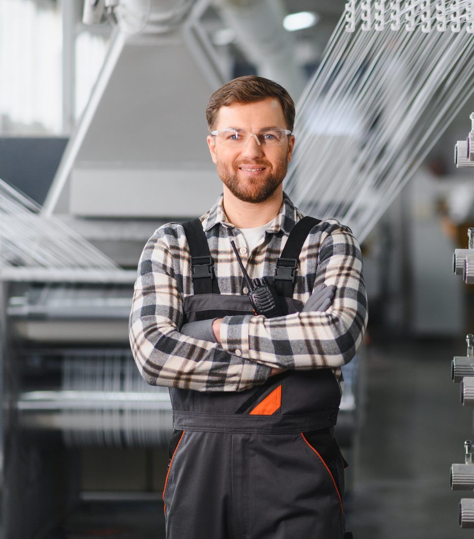 A smiling business owner in safety glasses and work overalls standing beside machinery in his factory