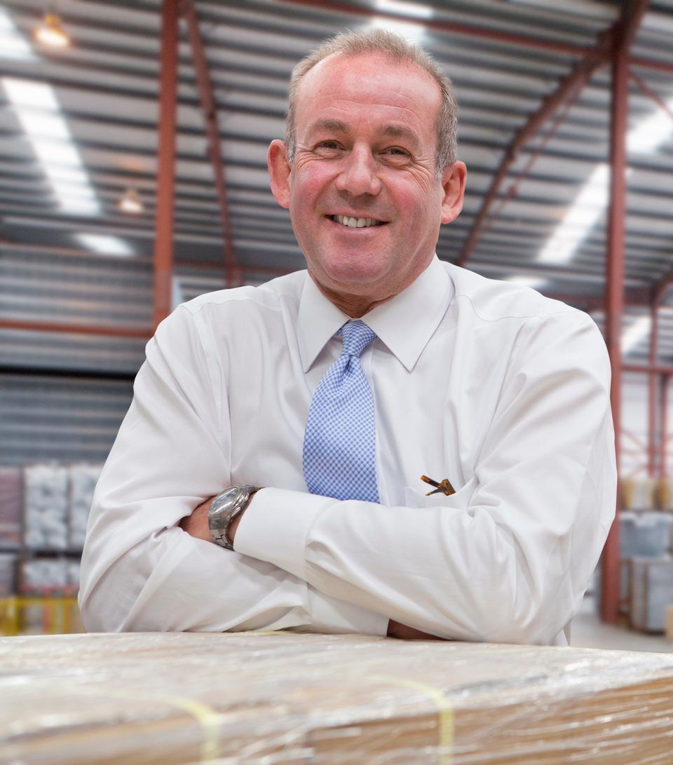 A smiling director in a shirt and tie leaning on a wrapped pallet in a large warehouse