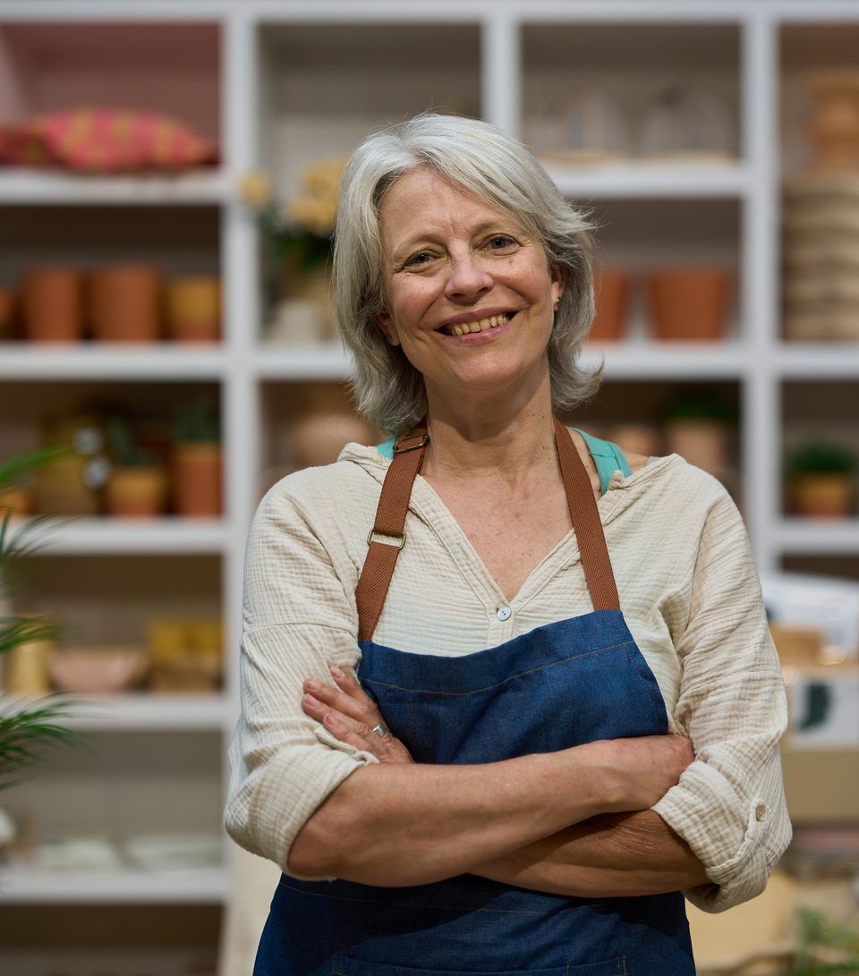 A smiling shop owner in a denim apron standing in front of shelves of pottery and homeware